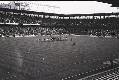 Vintage black-and-white stadium shot showing a midfield kickoff with players in formation. Crowded stands with tiered seating...