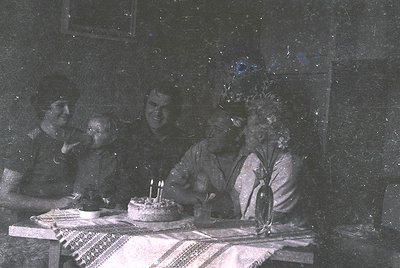 Vintage sepia-toned group portrait featuring five individuals seated around a table with a small cake and lit candles. Indoor...