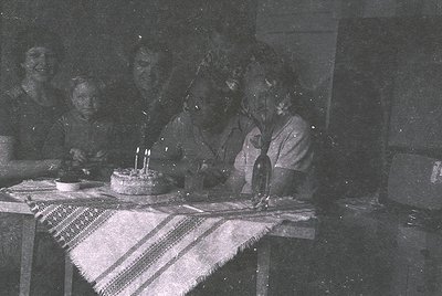 Vintage black-and-white indoor celebration featuring a woman blowing out candles on a simple round cake. Surrounding her are ...