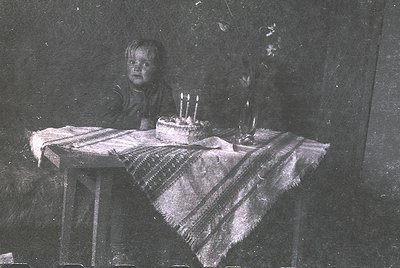 Vintage sepia-toned photo of a young child seated at a small wooden table covered with a striped cloth, blowing out birthday ...
