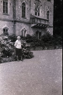 Black-and-white portrait of an elderly woman standing beside a Gothic Revival-style stone building with pointed arches and wr...