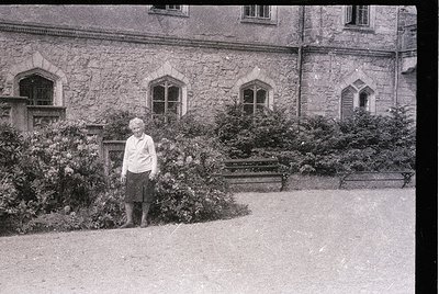Vintage black-and-white portrait of an elderly woman standing beside a stone building with arched windows, surrounded by dens...