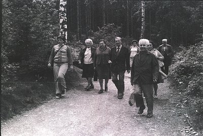 Group of seven adults walking on a dirt path through a forested area, 1970s. Casual attire suggests a leisurely hike or natur...