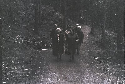 Black-and-white group hiking on a snow-dusted forest trail, likely mid-20th century. Six adults in winter coats and hats, one...