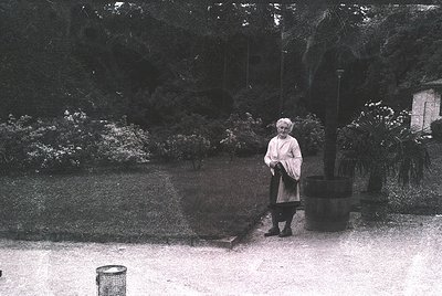A mid-20th century woman in a light coat and dark skirt stands in a landscaped garden, holding a handbag. Lush greenery, incl...