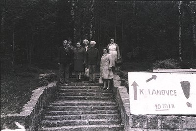 Black-and-white group photo on stone steps leading to a hand-painted wooden sign reading *"K LANOVCE 10 min"* in Czech, indic...
