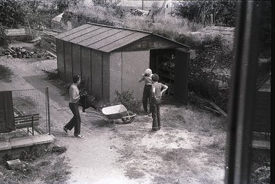 Mid-20th-century backyard scene with two men in casual workwear (bell-bottoms, button-ups) near a corrugated metal shed. One ...