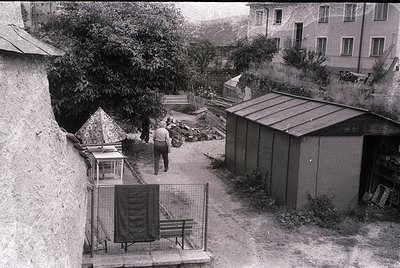 Vintage courtyard scene with concrete walls, metal fencing, and utilitarian structures. A man in mid-step walks toward a smal...