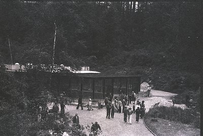 A vintage black-and-white photo of a forested hillside resort with a wooden pavilion and open-air seating. Mid-20th century a...
