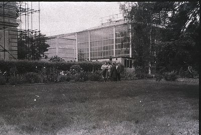 Mid-20th century botanical conservatory with large glass panels and scaffolding, likely 1950s-60s. Group of people in formal ...
