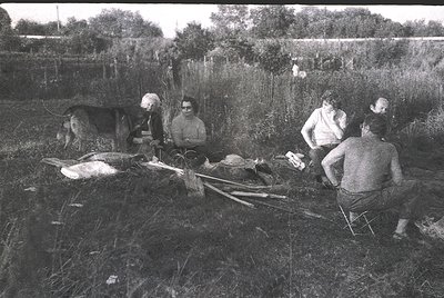 Mid-20th century rural gathering: group of five seated around an open-air meal in a grassy field, with a butchered animal car...