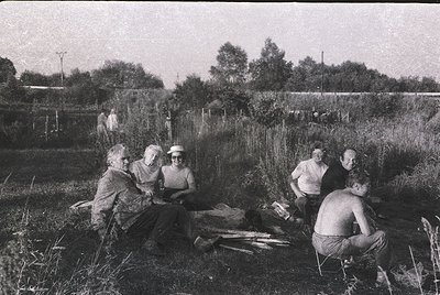 Five individuals seated in rural field, mid-20th century. Group includes three women in dresses and two men in shorts, posing...
