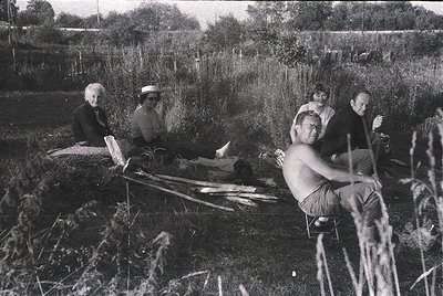 Group of five men in rural setting, mid-20th century. One man seated on logs, reading; others seated/leaning on natural mater...