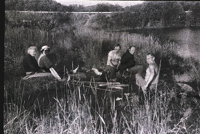 Five individuals relax by a shallow lake in a natural setting, mid-20th century. Two seated on logs, one reclining on grass, ...