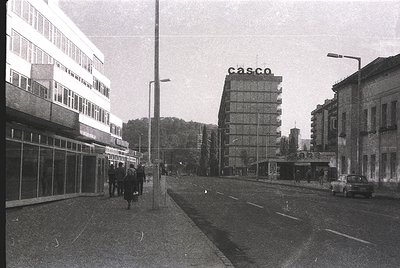 Mid-century urban street scene featuring Brutalist architecture. Prominent white concrete building with "casco" signage, like...