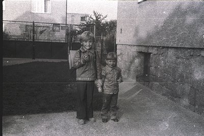 Two children pose in mid-20th-century urban setting, likely Eastern Europe. Boy on left wears a jacket with a patch, girl in ...