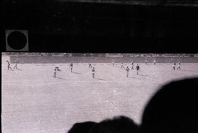 Vintage black-and-white shot of a soccer match in progress, likely mid-20th century. Players in striped jerseys and shorts en...