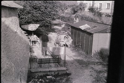A mid-20th century courtyard scene featuring a small group of people in casual 1960s attire. A man in a suit stands near a ga...