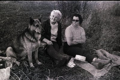 Black-and-white snapshot of two women and a German Shepherd in a rural setting, likely 1960s–1970s. Elderly woman in a cardig...