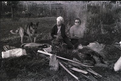 Two individuals seated outdoors on grass beside rustic tools and a dog, likely mid-20th century. The older woman wears a dark...