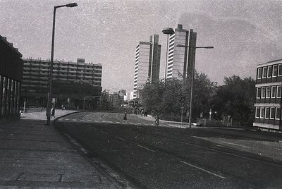 Mid-century urban street scene featuring Brutalist architecture: concrete high-rises with exposed block detailing flank a wid...
