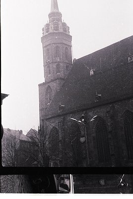 Vintage black-and-white photo of a Gothic-style church tower with ornate brickwork and a pointed spire, flanked by bare trees...