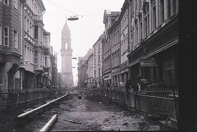 Bomb-damaged street in central European city, likely post-WWII. Ruined buildings flank a debris-strewn thoroughfare, with a s...