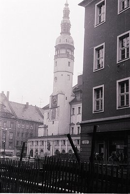 Historic European town square featuring a tall, white Baroque-style church tower with a spire and clock, flanked by multi-sto...