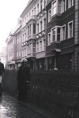 Mid-20th century European street scene featuring classic brick buildings with ornate balconies and large windows. A woman in ...