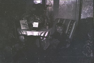 Vintage sepia-toned interior shot of a child seated on ornate, patterned upholstery beside a small table. Table displays a fr...