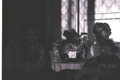 Vintage black-and-white interior shot of a seated woman in a patterned dress, holding a cigarette holder. Center table displa...