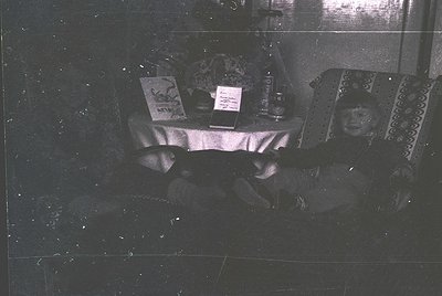 Vintage black-and-white interior shot of a dimly lit room with a low table covered in patterned cloth. Two individuals seated...
