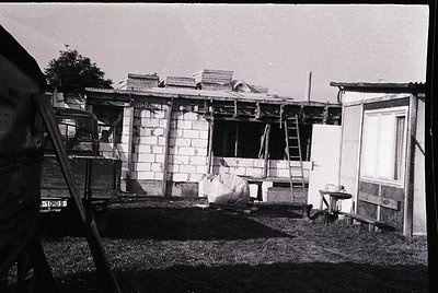 Mid-20th century mobile home under construction, brick exterior with wooden framing visible. Ladder leans against structure; ...