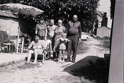 Family portrait from the 1960s–70s, likely Eastern Europe. Five adults and a child pose outdoors near a shaded umbrella and v...
