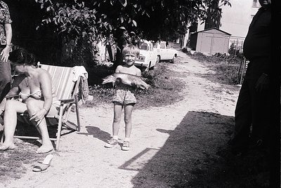 Vintage black-and-white snapshot of a mid-20th-century backyard scene. A young girl in floral shorts and sandals stands holdi...