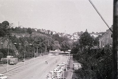 Vintage black-and-white street scene with parked vintage cars, likely 1960s–1970s. Urban landscape featuring a mix of residen...