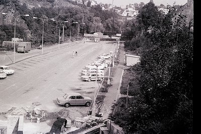 Aerial view of a mid-20th-century seaside parking lot with parked cars lining both sides. Concrete barriers and utility poles...