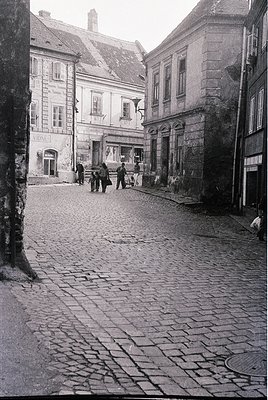 Cobblestone street in a European town with mid-20th century architecture—stone buildings with arched doorways and shuttered w...