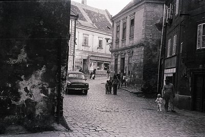 Mid-20th century European street scene with cobblestone pavement, vintage car (likely or ), and stone buildings. People dress...