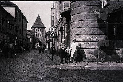 Mid-20th century European street scene with cobblestone pavement, featuring a woman in a long dress holding a child’s hand. D...