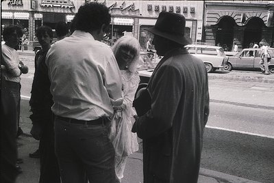 Black-and-white street scene featuring three central figures: a woman in a long, sheer wedding gown adjusting her veil, flank...