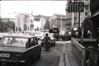 Urban street scene with mid-20th century vehicles, including a motorcycle and vintage cars. Pedestrians cross near a pedestri...