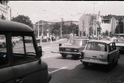 Vintage urban street scene with mid-20th century Soviet-era cars (Lada/ZAZ) and a bus, likely Eastern Bloc. Pedestrians cross...
