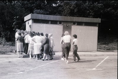 Mid-20th century group photo outside a utilitarian concrete block changing room. Women in 1960s-70s fashion—pleated skirts, b...