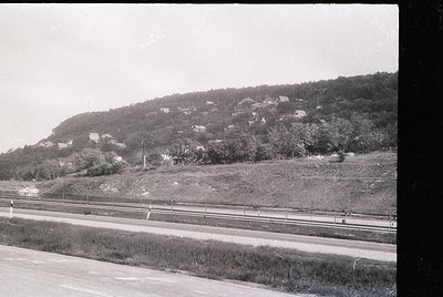 Black-and-white rural landscape featuring a winding road bordered by a double railway track. Dense, terraced hillside settlem...