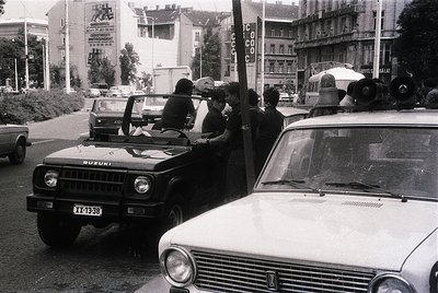 Black-and-white street scene featuring a Suzuki off-road vehicle with a group of people standing on its roof, likely during a...