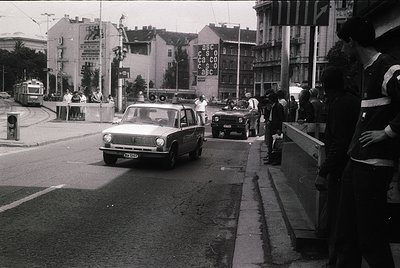 Vintage urban street scene featuring a 1970s-era sedan driving past a pedestrian bridge. Crowd on sidewalk, some wearing high...