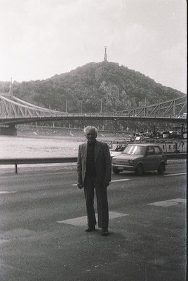 Mid-20th century black-and-white photo: Man in 1960s-era suit stands on a paved plaza beside a bridge, with a prominent hillt...
