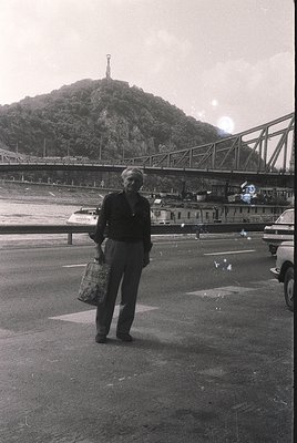Mid-20th century black-and-white photo of a man standing near a bridge with a statue atop a hill in the background. He holds ...