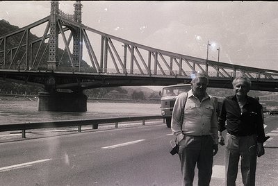 Black-and-white shot of two men in 1960s-era attire standing beside a multi-lane road under the **Freedom Bridge (Liberty Bri...